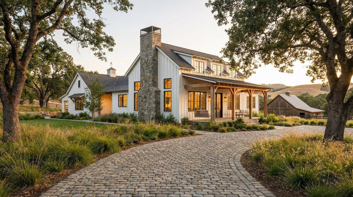 White farmhouse with a stone chimney and a curving cobblestone driveway under oak trees