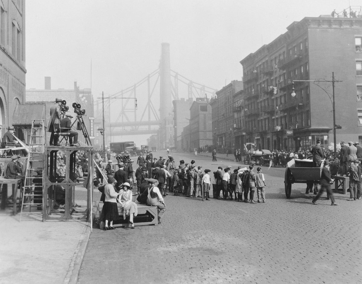 Historic black-and-white photo of a New York cobblestone street crowded with people, with a bridge in the background